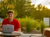 A young man sits with a laptop open and smiles